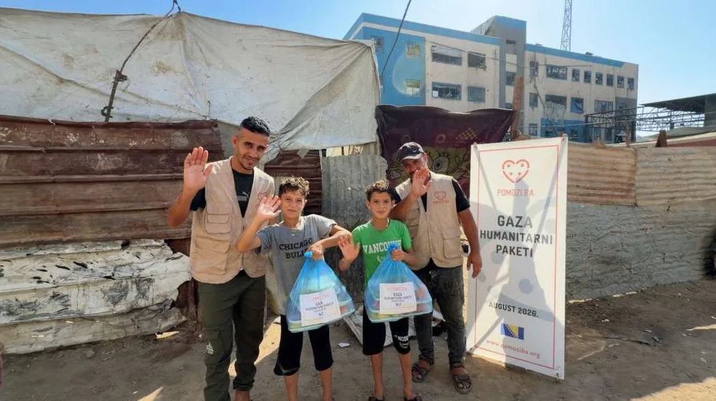 Dvoje muškaraca i dvoje dječaka mašu na kameri, držeći pakete s humanitarnom pomoći u Gazi.
