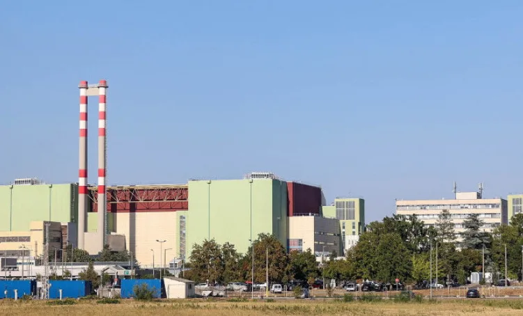 Industrijski kompleks sa dva visoka dimnjaka na lijevoj strani, zgradama različitih boja i zelenim drvećem ispred plavog neba.