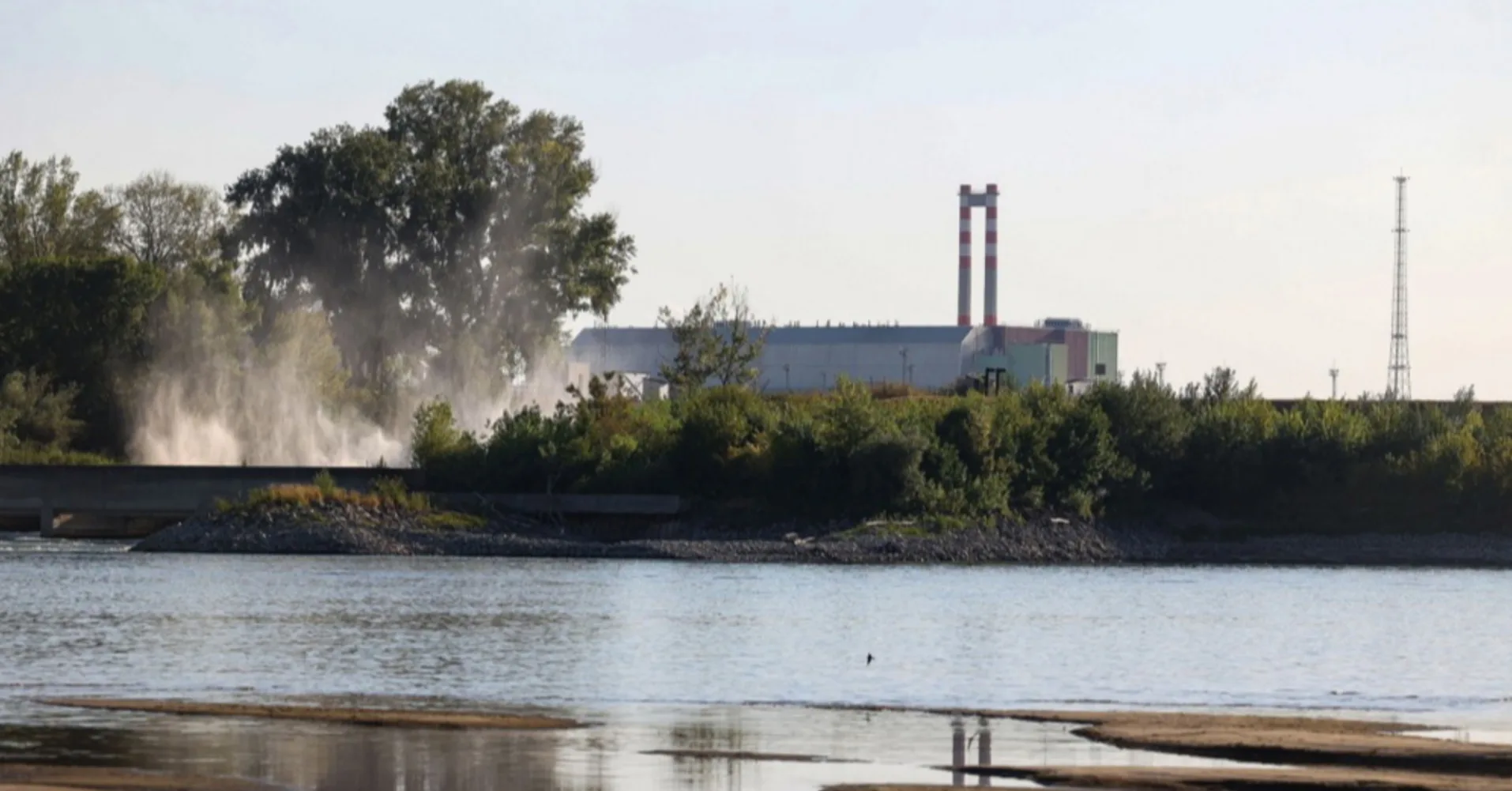 Industrijski objekat sa visokim crveno-bijelim dimnjakom, smješten pored bujne zelene vegetacije i rijeke, uz maglovitu izmaglicu koja se širi iznad vodene površine i drveća.