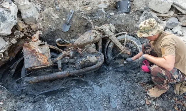 Čovjek u maskirnoj uniformi i šeširu sa šarama pregleda iskopani zarđali motocikl iz blata.