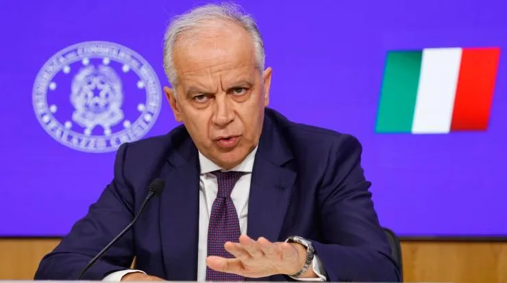 Muškarac u tamnom odijelu i ljubičastoj kravati govori ispred plave pozadine sa znakom Italije i zastavom Italije.