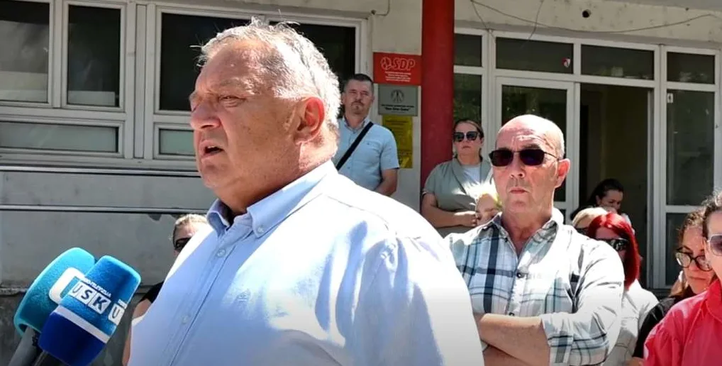 Čovjek u plavoj košulji daje izjavu ispred zgrade, držeći se za mikrofon, sa grupom ljudi u pozadini.