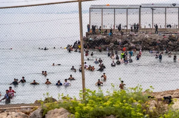 Grupa ljudi pliva u plitkom moru, neki stoje u vodi do pojasa, dok drugi plivaju dalje od obale. U pozadini se vidi kameni nasip sa visokom ogradom, iza koje stoje ljudi koji izgledaju kao granični službenici. Mnogi ljudi su u vodi, neki izgledaju kao da pokušavaju da pređu granicu, dok drugi provode vrijeme u moru.