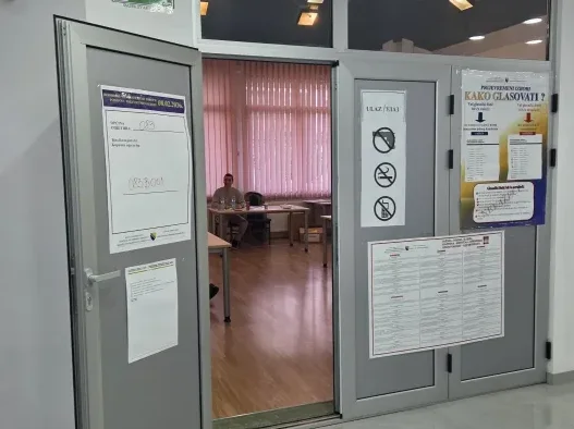 Fotografija prikazuje ulaz u prostoriju izborne jedinice u Bosni i Hercegovini. Na vratima su istaknuti informativni plakati o izbornom procesu, uključujući upute za glasanje i popis birača. U pozadini se vidi unutrašnjost prostorije sa stolovima i stolicama, te osoba koja sjedi za stolom, vjerovatno član biračkog odbora. Prostorija je osvijetljena, a prozori su prekriveni roletnama.