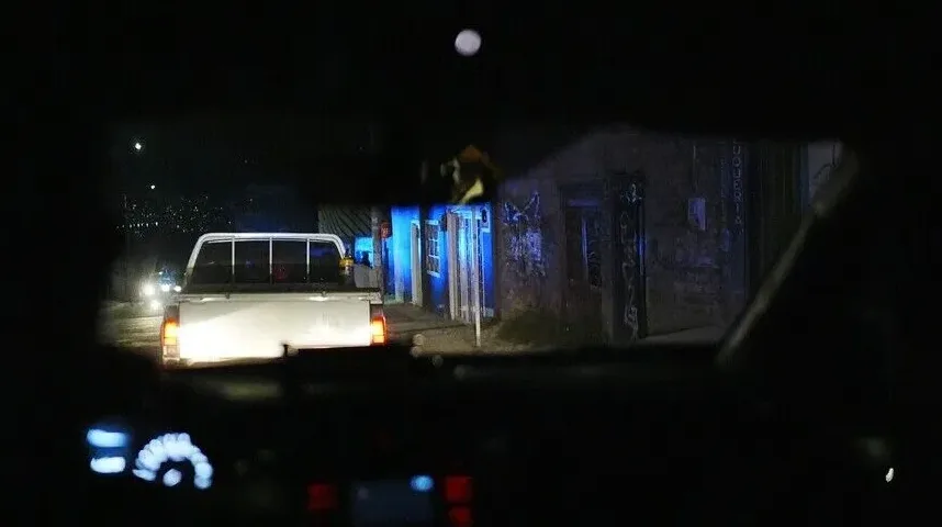 Bijeli kamion sa uključenim kočnicama zaustavljen na mračnoj ulici noću, osvijetljen plavim svjetlima zgrade u pozadini.