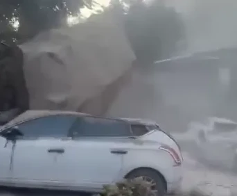 Bijeli automobil parkiran ispred kuće prekrivene prašinom i ruševinama od klizišta, uz vidljive ostatke zemlje i kamena na vrhu