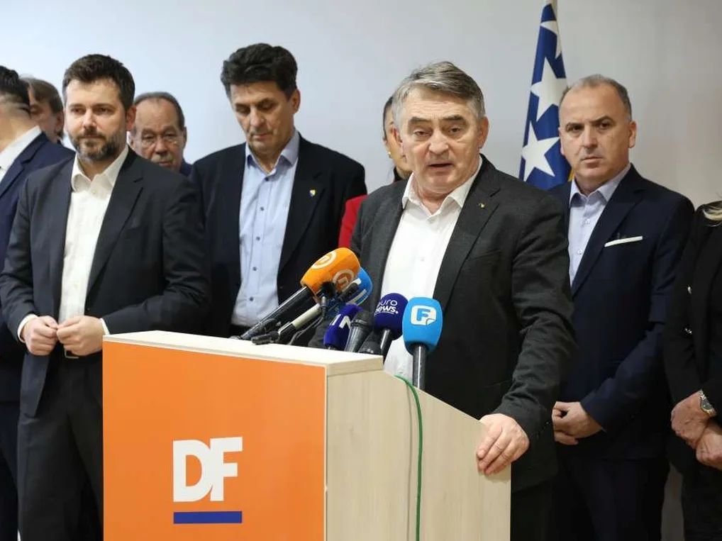Članovi Demokratske fronte okupljeni oko podijuma s mikrofonima, sa zastavom Bosne i Hercegovine u pozadini.