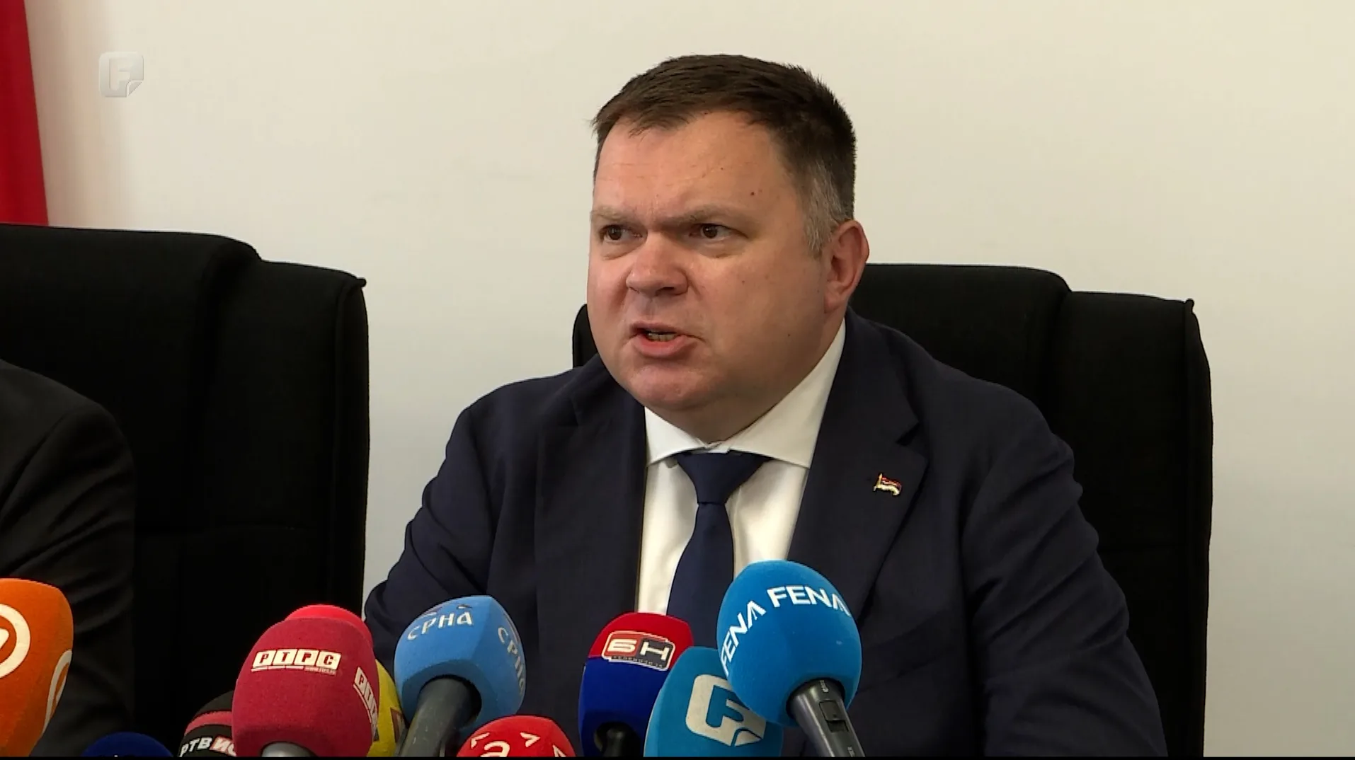 Političar u tamnom odijelu i plavoj kravati sjedi iza stola s brojnim mikrofonima medijskih kuća ispred sebe i govori.