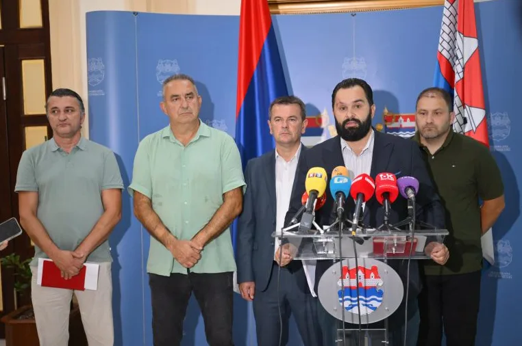 Četiri muškarca stoje iza govornice s mikrofonima, od kojih jedan drži govor. Iza njih se nalaze zastave i plavi zid s logotipima.