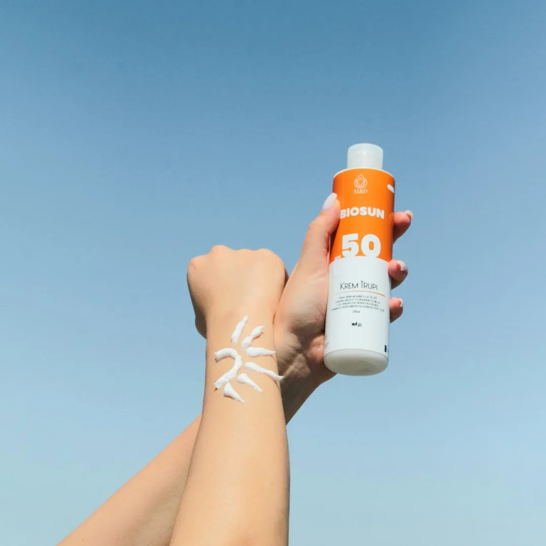 Ruka koja drži bocu kreme za sunčanje Biosun SPF 50 s nacrtanim suncem na podlaktici.
