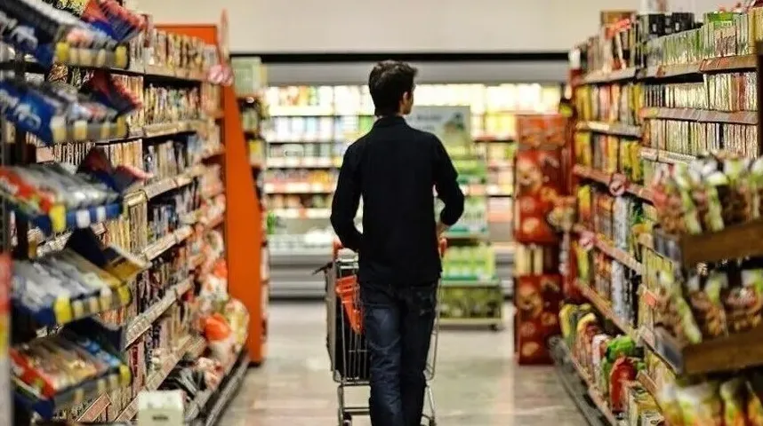 Mladić u tamnoj jakni gura kolica punog namirnica kroz prolaz u supermarketu, okružen policama sa robom.