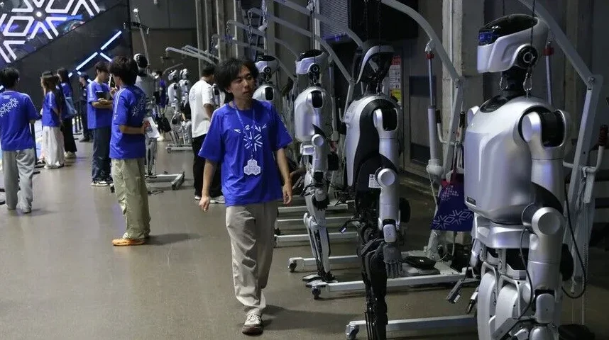 Grupa ljudi, među kojima su neki u plavim majicama, stoje pored reda humanoidnih robota na izložbi ili sajmu tehnologije.