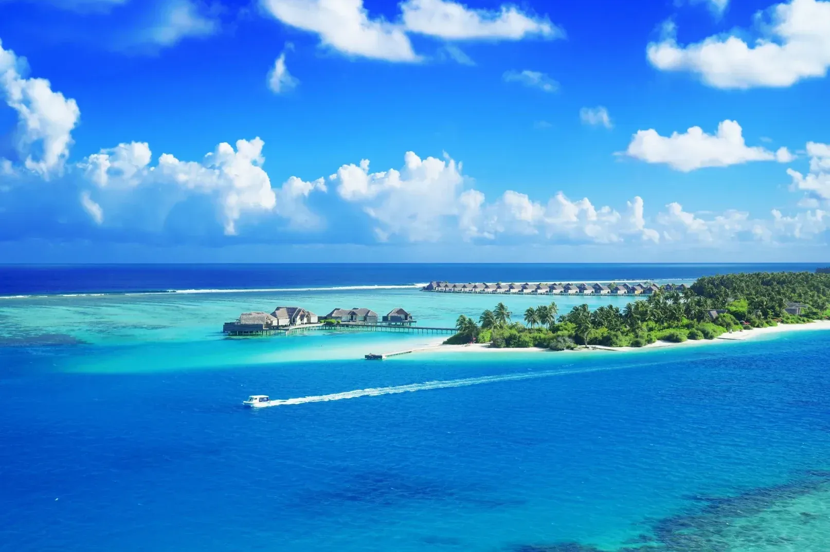 Pogled na tirkizno plavo more, pješčani sprud, tropsku vegetaciju i redove bungalova na stubovima iznad vode na Maldivima.