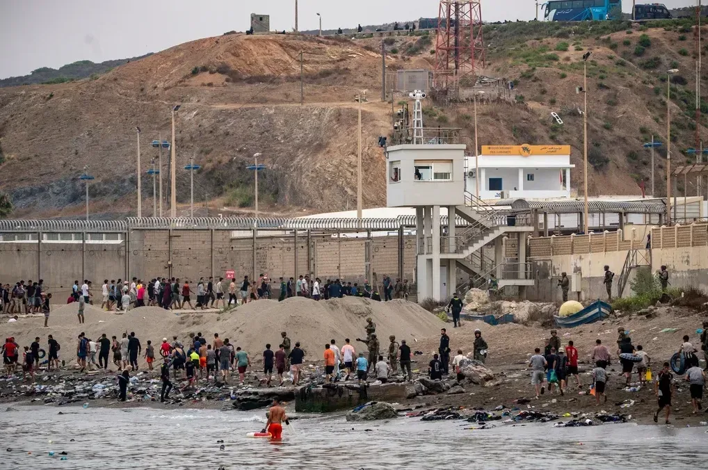Grupa migranata okupljena uz obalu blizu visoke ograde i nadzorne kule, sa pripadnicima sigurnosti.