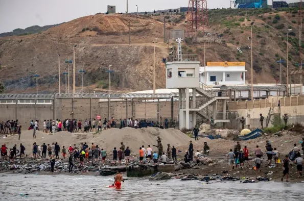 Grupa migranata se okuplja blizu visoke ograde i nadzorne kule sa naoružanim stražarima.