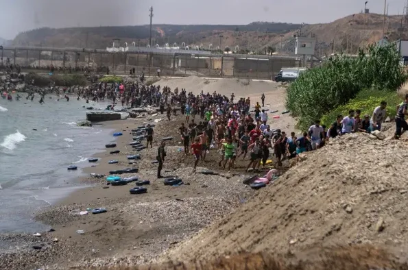 Velika grupa migranata trči prema obali, neki ulaze u more, dok drugi napuštaju vodu. U pozadini se vidi ograda i brdovit teren.