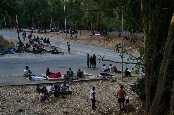 Grupa migranata odmara se pored puta, neki leže na zemlji, drugi sjede, okruženi drvećem i krhotinama.