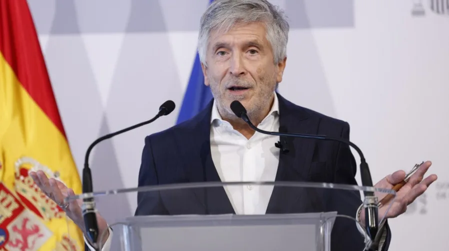 Čovjek sa sijedom kosom i bradom, obučen u tamni sako i bijelu košulju, drži olovku u ruci dok govori za govornicu.
