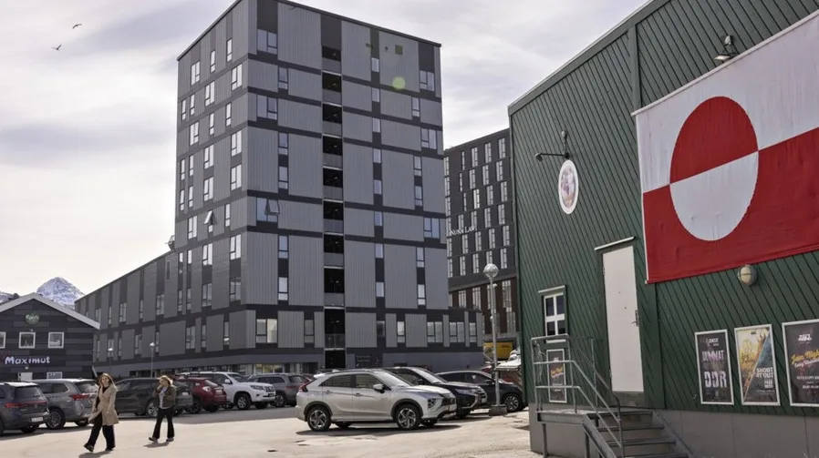 Moderna stambena zgrada sa sivom fasadom i brojnim prozorima, parkiranim automobilima ispred, te zelenom zgradom sa istaknutom zastavom Grenlanda.