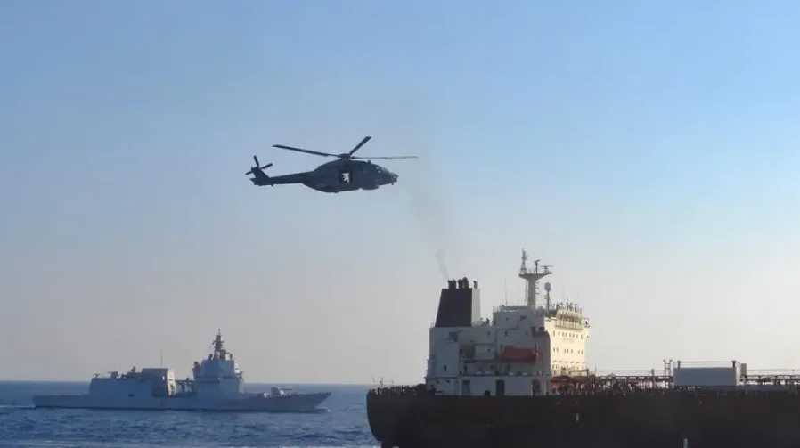 Mornarički helikopter leti iznad mora, sa ratnim brodom u pozadini i teretnim brodom u prvom planu.
