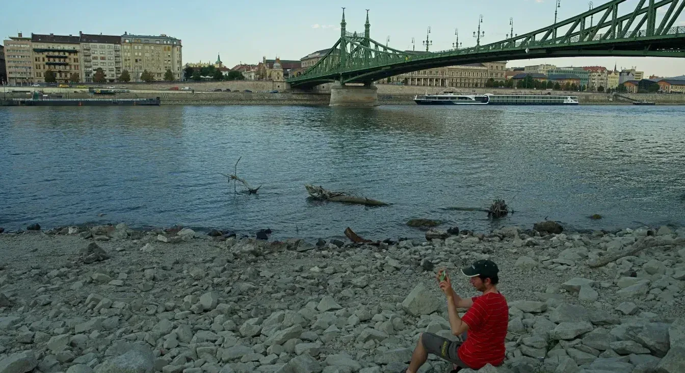 Čovjek sa šeširom sjedi na šljunkovitoj obali Dunava i fotografiše zelenim mobilnim telefonom. U pozadini se vidi zeleni most i zgrade Budimpešte.