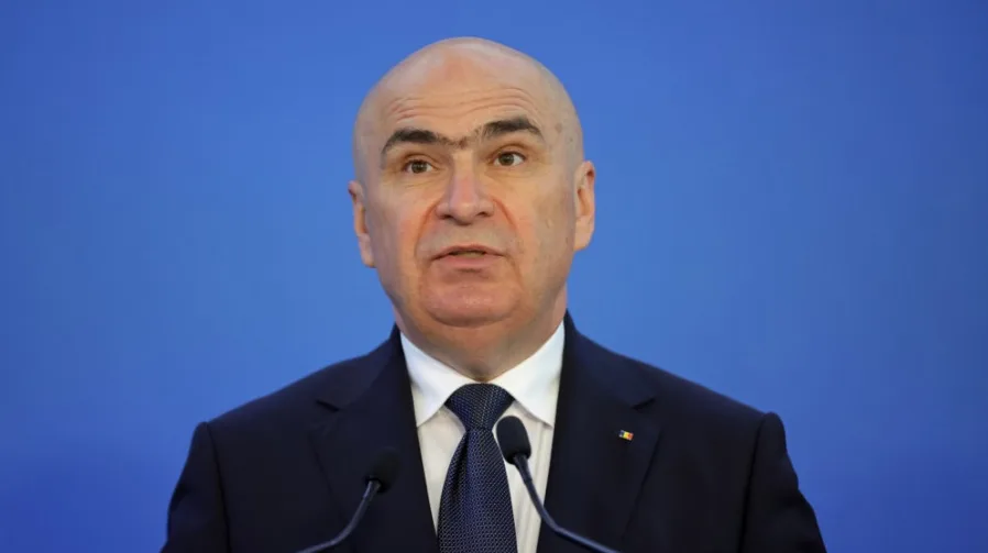 Portret ćelavog muškarca u tamnom odijelu sa tamnoplavom kravatom, koji stoji ispred plave pozadine i govori za govornicu.