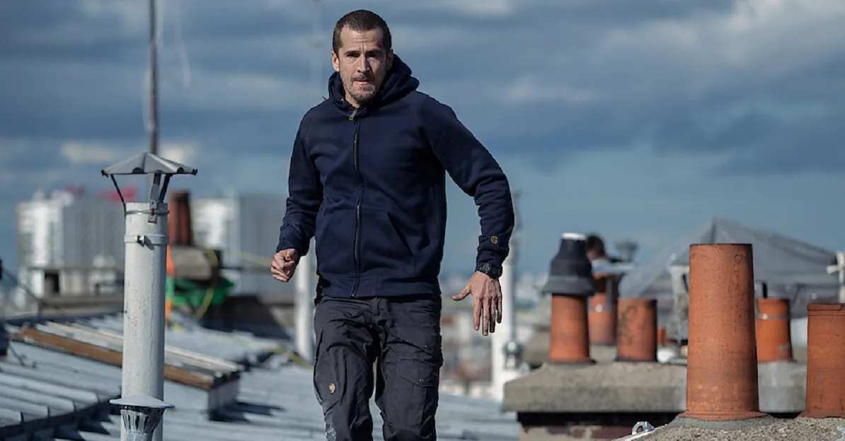 Muškarac u tamnoj dukserici i cargo pantalonama trči po krovu zgrade, okružen dimnjacima i urbanim pejzažem u pozadini.