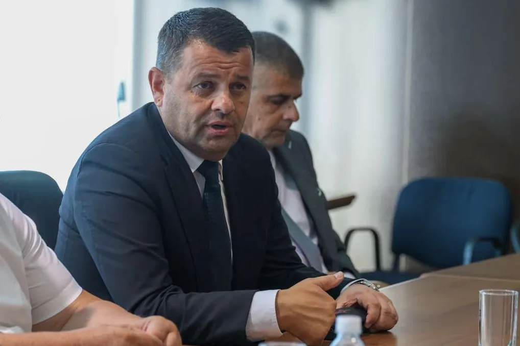 Muskarac u crnom odijelu sa bijelom kosuljom i tamnom kravatom, sjedi za drvenim stolom i govori, pogled mu je usmjeren lijevo. U pozadini se naziru jos dva muskarca i plava stolica.