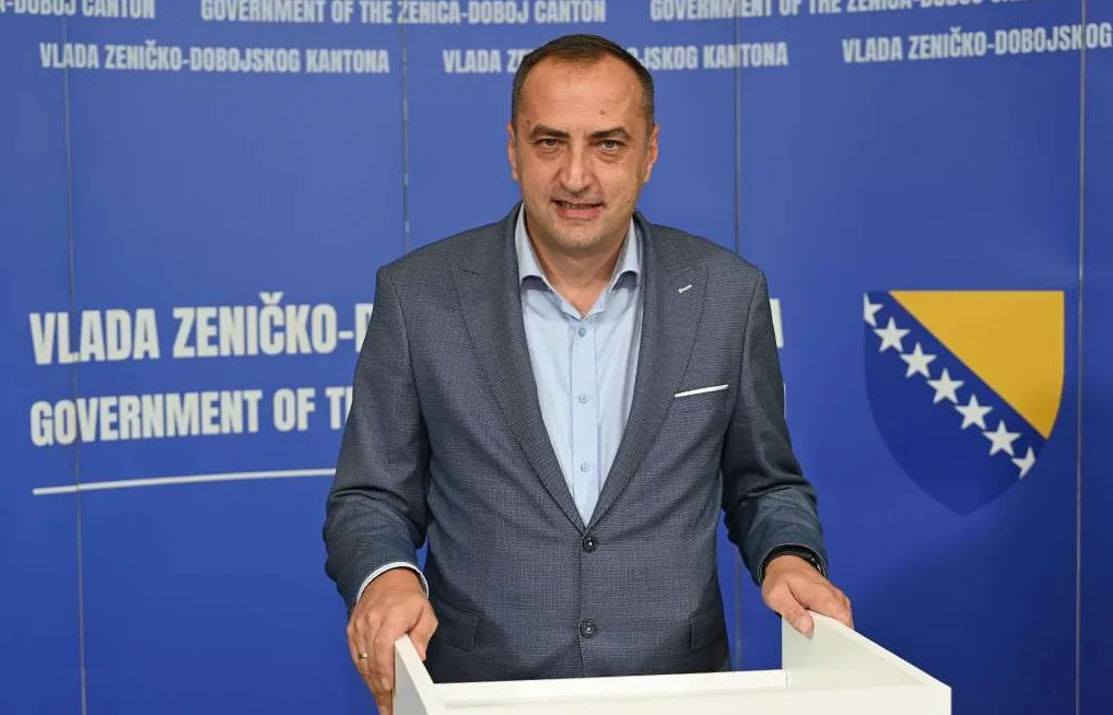 Portret muškarca u tamnom odijelu i plavoj košulji, koji stoji ispred plave pozadine sa logom Vlade Zeničko-Dobojskog kantona i zastavom Bosne i Hercegovine.