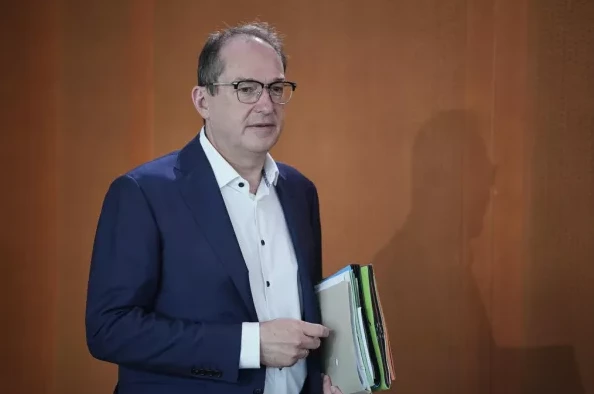 Muškarac u tamnoplavom odijelu i bijeloj košulji sa naočalama, drži fascikle u ruci.