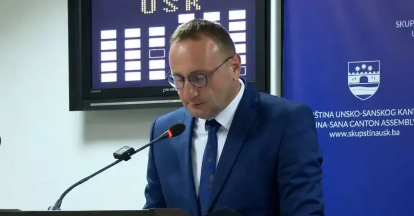 Muškarac u tamnoplavom odijelu i plavoj kravati, naočarima, govori za govornicom sa mikrofonom.