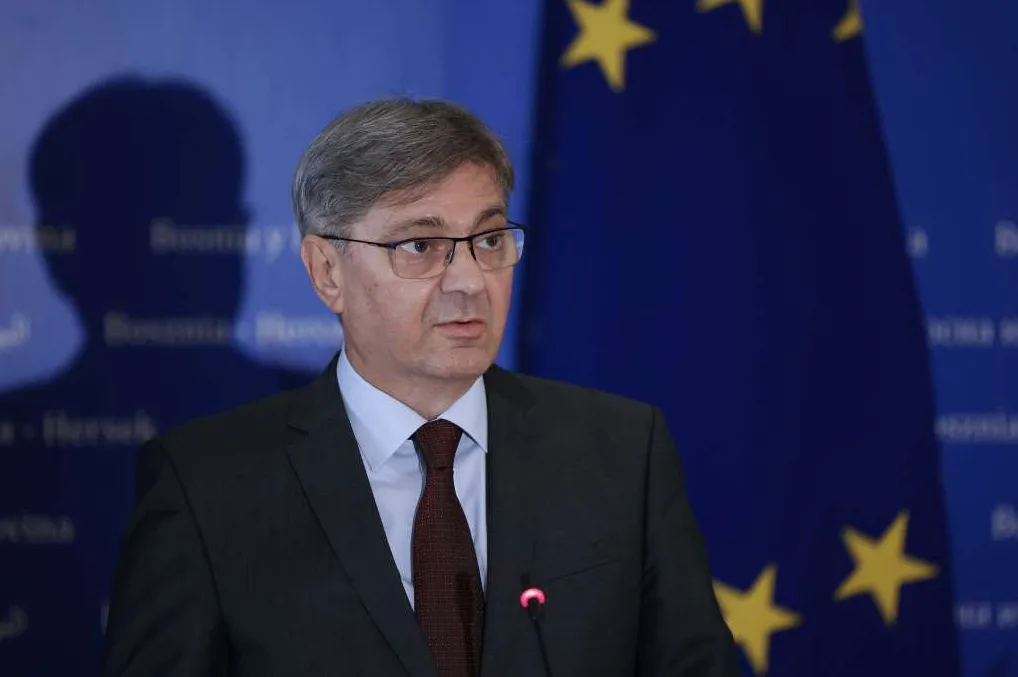 Muškarac u tamnom odijelu i crvenoj kravati, nosi naočale i stoji ispred plave zastave Evropske unije.