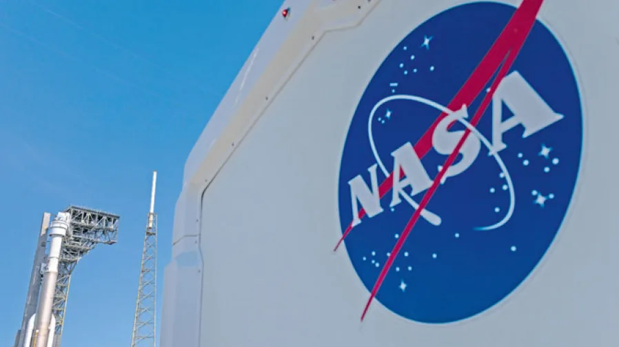 Logotip Nacionalne vazduhoplovne i svemirske administracije (NASA) na bijelom vozilu, s raketnom rampom u pozadini pod plavim nebom.
