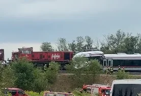 Dva voza su se sudarila na pruzi, jedan je crveni teretni voz, a drugi bijeli putnički voz. Vatrogasci u žutim uniformama su na mjestu događaja.