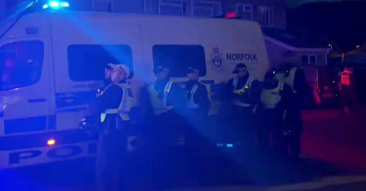 Grupa policajaca u uniformama i sa prslucima visoke vidljivosti stoji pored policijskog kombija sa ukljucenim plavim rotacijama. Na kombiju se vidi natpis "NORFOLK POLICE".