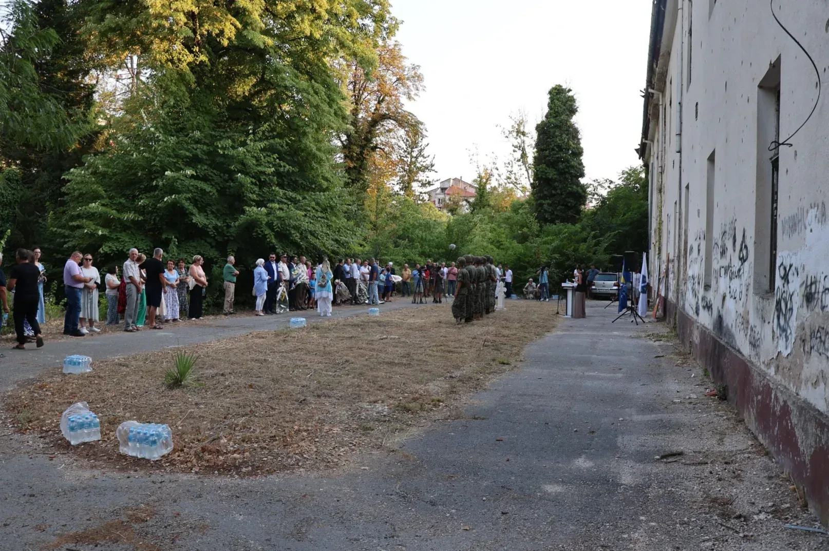 Grupa ljudi stoji u redu ispred devastirane zgrade, poredani uz drveće, dok se na drugoj strani primećuje postolje s govornicom i zvučnicima.