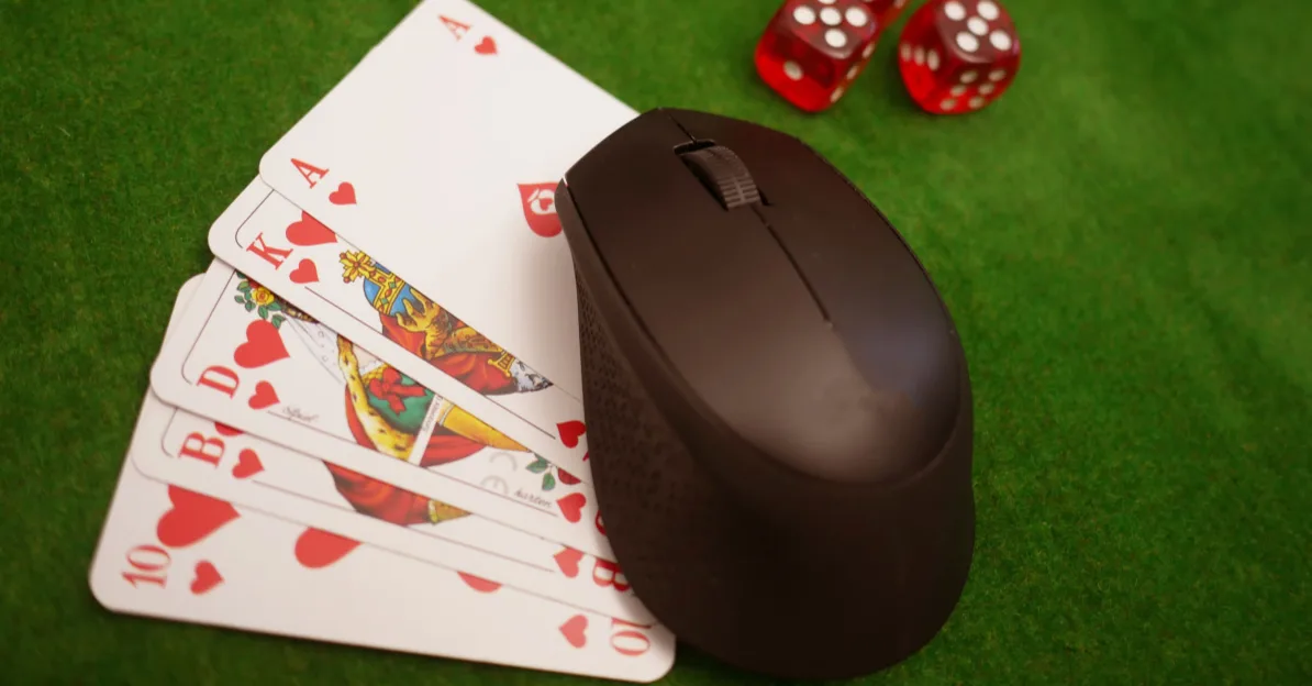 Kompjuterski miš leži na poker kartama (As, Kralj, Dama, Devetka, Desetka) sa srcem kao bojom, pored crvenih kocki na zelenoj podlozi za kockanje.