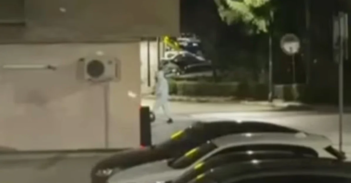 Osoba obučena u bijelo odijelo kreće se noću ispred zgrade sa parkiranim automobilima.
