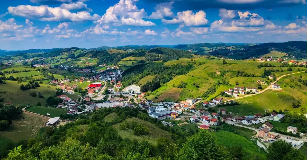 Panoramski pogled na zeleno selo smješteno u dolini okruženoj brežuljcima prekrivenim šumom i bujnom vegetacijom, s bijelim oblacima na plavom nebu.