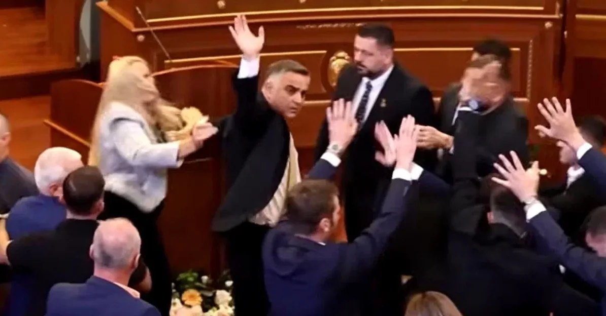 Muškarac u crnom odijelu podiže ruku, okružen drugim osobama u sličnim odijelima, u prostoriji nalik parlamentarnoj dvorani.