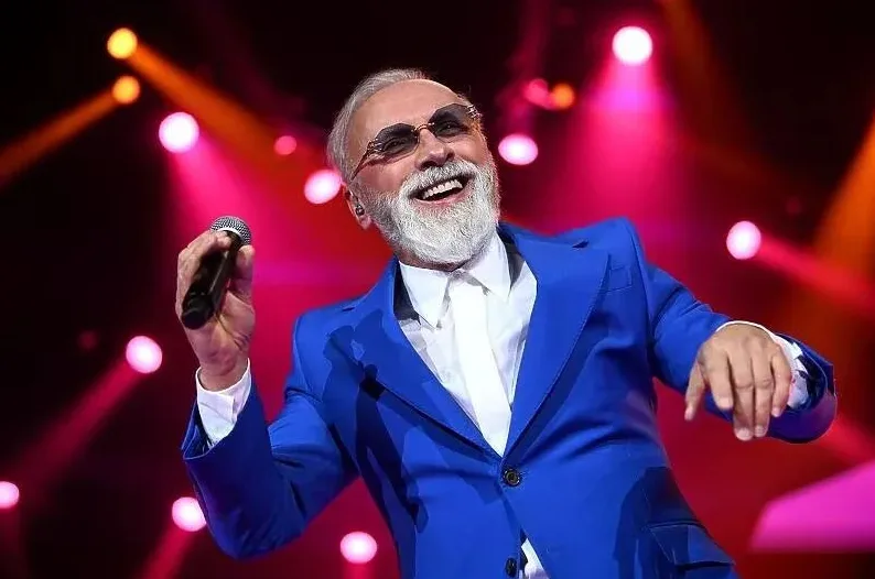 Muzičar sa bijelom bradom i kosom, obučen u plavo odijelo i bijelu košulju, drži mikrofon i pjeva na sceni obasjanoj ružičastim i narandžastim reflektorima.