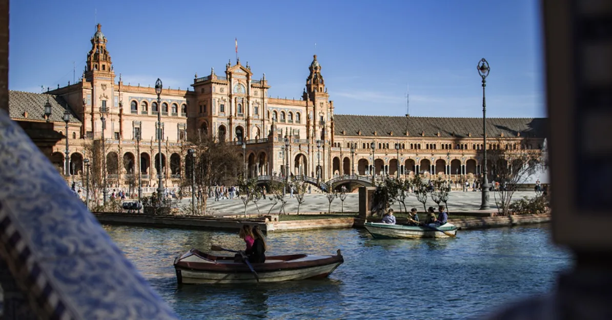Turisti uživaju u vožnji čamcem po kanalu ispred raskošne arhitekture Trga Španije (Plaza de España) u Sevilji.