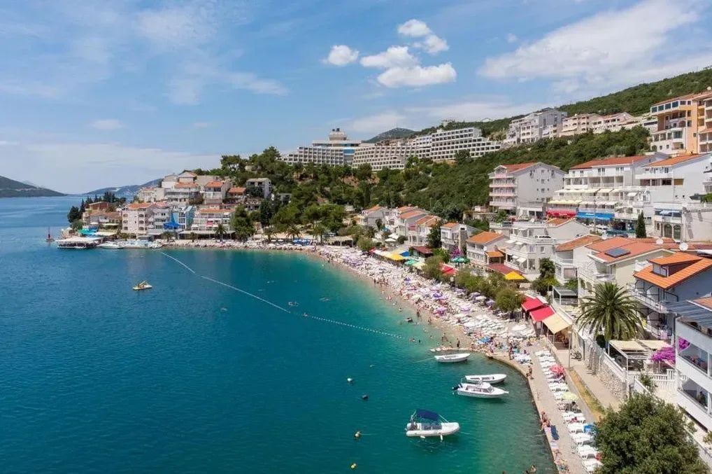 Aerijalni pogled na živopisnu plažu sa mnogo posjetilaca, ležaljkama i suncobranima, okruženu urbanim naseljem i hotelskim kompleksima na padini brda, te azurno plavim morem sa nekoliko brodica na njemu.