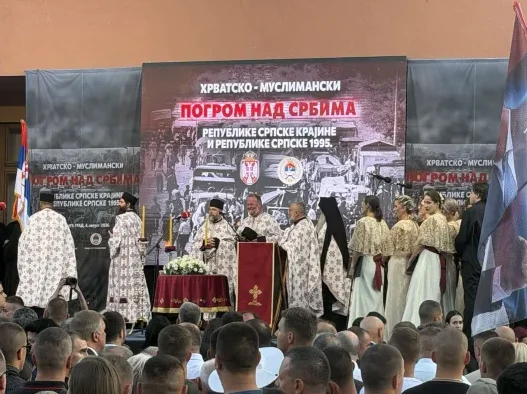 Sveštenici SPC u svečanim odorama stoje ispred scene na kojoj je natpis "Hrvatsko-muslimanski pogrom nad Srbima u Republici Srpskoj Krajini i Republici Srpskoj 1995.", dok publika stoji i posmatra.