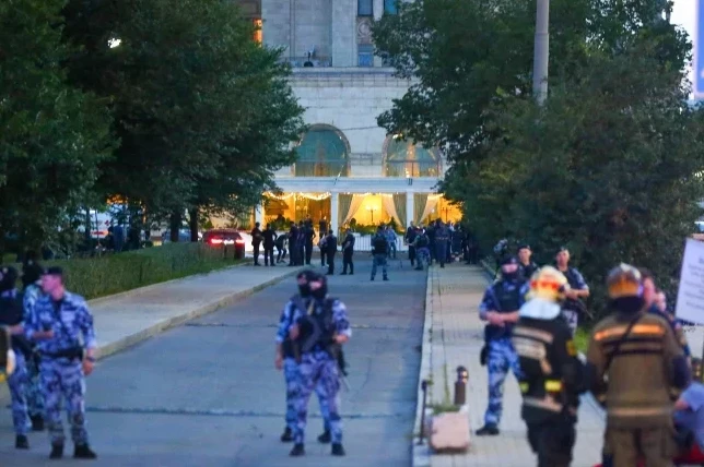 Snimak prikazuje grupu uniformisanih policajaca i vatrogasaca ispred velike zgrade sa osvijetljenim ulazom.