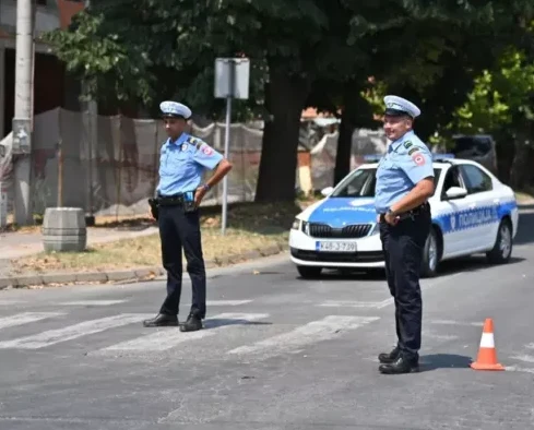 Dva policajca u uniformi stoje na pješačkom prelazu, na ulici, pored policijskog automobila.