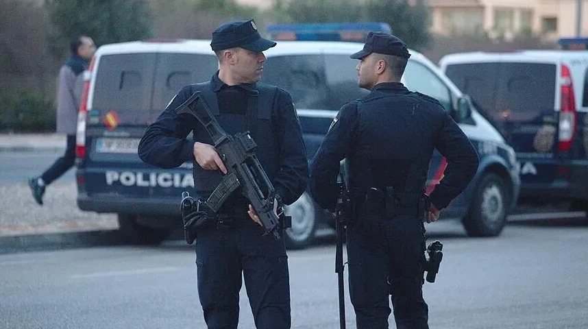 Dva policajca u uniformi stoje na ulici, jedan od njih drži automatsku pušku, a iza njih se vide policijska vozila.