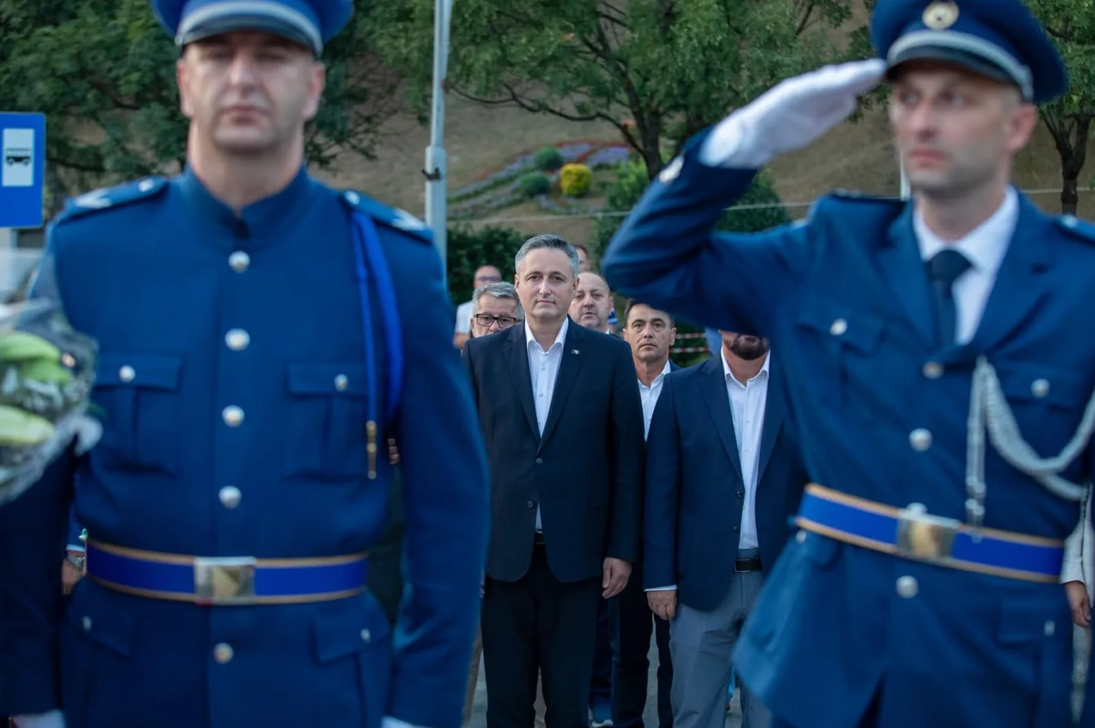 Dva policajca u plavim uniformama, jedan s lijeve strane u punom fokusu, a drugi desne strane vrši počasni salut. Iza njih, u pozadini, stoji grupa muškaraca u formalnim odijelima, sa istaknutim čovjekom u tamnom odijelu u sredini.