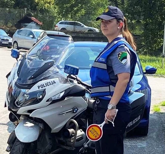 Policajka u uniformi i fluorescentnom prsluku stoji pored policijskog motocikla držeći STOP znak.
