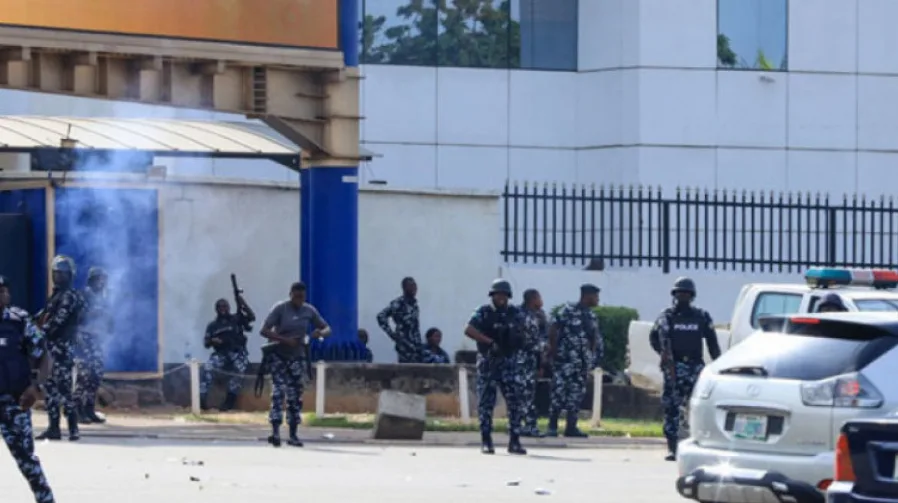 Policajci u uniformama, neki sa oružjem, okružuju zgradu dok suzavac ispušta plavi dim.